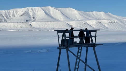 Researchers installing weather station equipment on platform in Svalbard, with a snowy mountainous landscape and blue skies behind