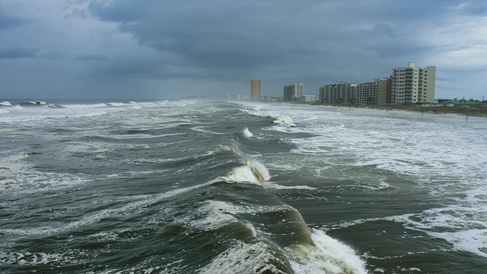 Looking down coastline with large white waves and stormy skies as hurricane approaches coastline with skyscrapers on shore