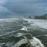 Looking down coastline with large white waves and stormy skies as hurricane approaches coastline with skyscrapers on shore